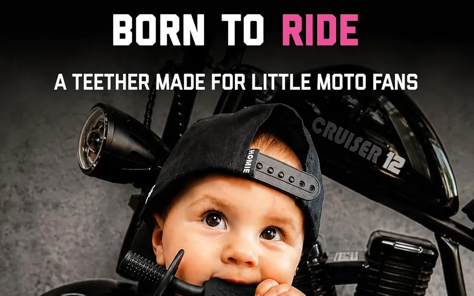 A baby in a cap chewing a motocross-style teether in front of a motorcycle